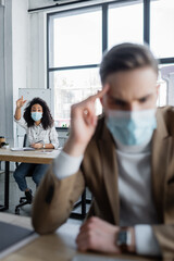 Naklejka premium african american businesswoman in protective mask waving hand behind colleague suffering from headache on blurred foreground
