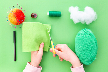 Instruction step by step, step one. The hands of a child in the process of sewing a toy from felt, a green one-eyed monster. Green background, flat lay. Hobbies, handicrafts, fine motor skills.