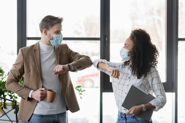 young multicultural managers in protective masks doing elbow greeting in office