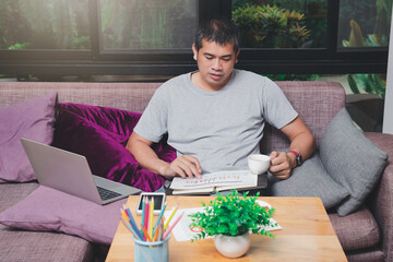 Asian man holding a cup of coffee and working while sitting on sofa in home office.