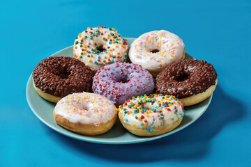 Bad food. Close up shot of various colourful round glazed donuts with sprinkles on the plate over blue background