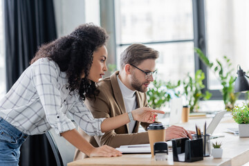 African american businesswoman pointing at laptop near colleague in office