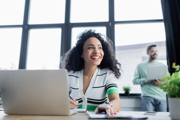 Cheerful african american businesswoman holding pen near notebook, laptop and clipboard on blurred foreground