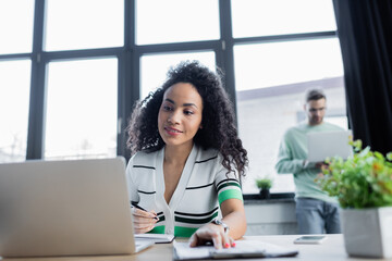 Smiling african american businesswoman working with clipboard and laptop on blurred foreground