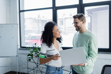 Smiling businessman with laptop looking at african american colleague in office