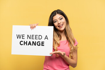 Young Russian girl isolated on yellow background holding a placard with text We Need a Change and  pointing it