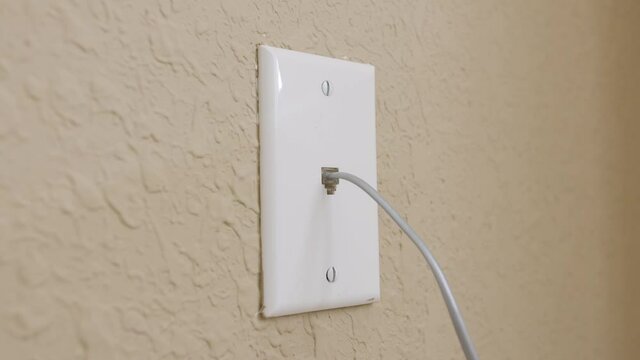 Male electrician plugging in landline telephone network cord into wall mounted jack plate.