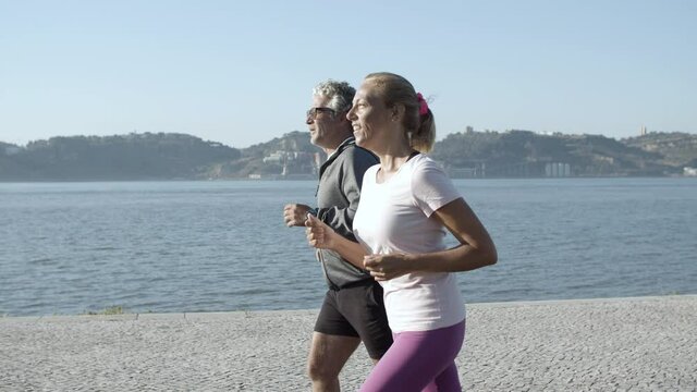 Active couple training, jogging along sea and smiling. Senior man and middle-aged woman running on nature together. Medium shot. Tracking camera. Healthy lifestyle and sport activity concept