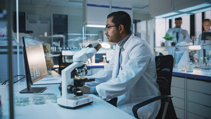 Medical Research Scientist Conducts DNA Experiments With Use of Microscope, Writes Results on Desktop Computer in Biological Science Laboratory. Diverse Multiethnic Lab with Engineers in White Coats.