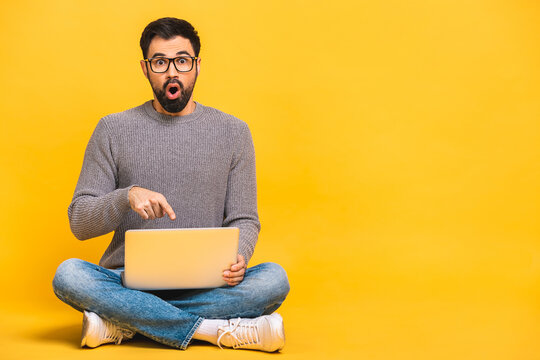 Young bearded man shocked surprised amazed with laptop computer. Funny image of young Caucasian male student model sitting on floor isolated on yellow background.