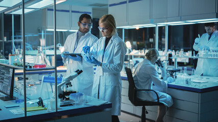 Obraz premium Female and Male Medical Research Scientists Have a Conversation While Conducting Experiments in Sample Test Tubes, Writing Analysis Results on a Tablet Computer. Modern Biological Science Laboratory.