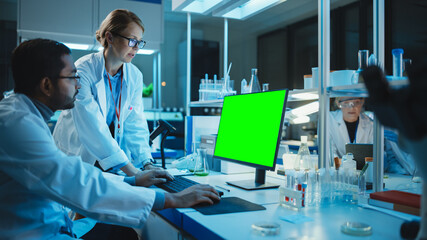 Female Research Scientist Has a Conversation with Bioengineer Next to a Desktop Computer with Green Screen Mock Up. They Look at Computer Template in a Modern Science Laboratory.