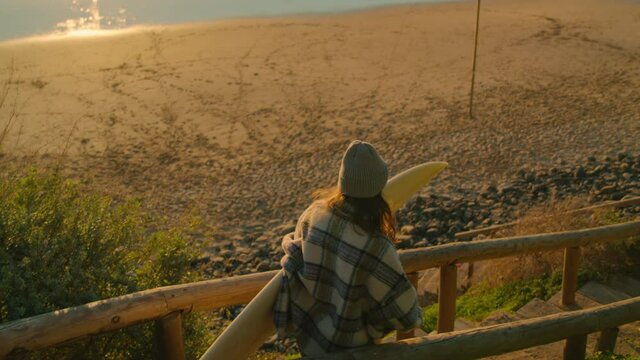Handheld slow motion video of young millennial woman in casual clothes walk down to epic surfing beach at sunset. Female surfer ready for surfing session. Cinematic vanlife surf lifestyle concept