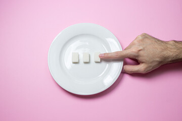 Sugar on a pink background. Three cubes of sugar on a white plate. The hand reaches for the sugar.