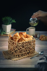 A traditional French croissants in a wooden basket, with hands pouring tea from a pot into a cup, black background with copy space.It's a buttery, flaky pastry fold with butter in multiple layers   
