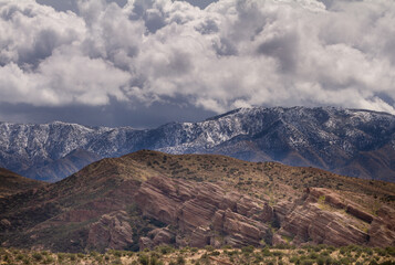 Layers of landscapes in the desert.  Green bushes in the foreground, angled rocks in the middle, and mountains covered with fresh snow.  All of this sits under moody skies. 