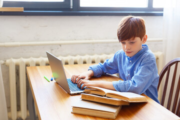 Teen boy doing homework online at home. He looks at the textbook and types a message with questions to the teacher.