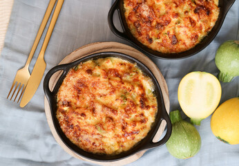 Top view of a traditional zucchini bake gratin in golden color of grilled grated cheese on top in a black round casserole, including its fresh colorful fruit with fork and spoon in background  