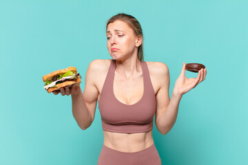 young pretty sport woman doubting or uncertain expression and holding a sandwich and a donut