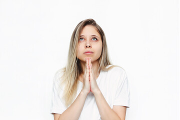 A young caucasian beautiful blonde woman in a white t-shirt prays with folded hands and looks up at the sky,  thanks, making wish, asking help or forgiveness isolated on white background