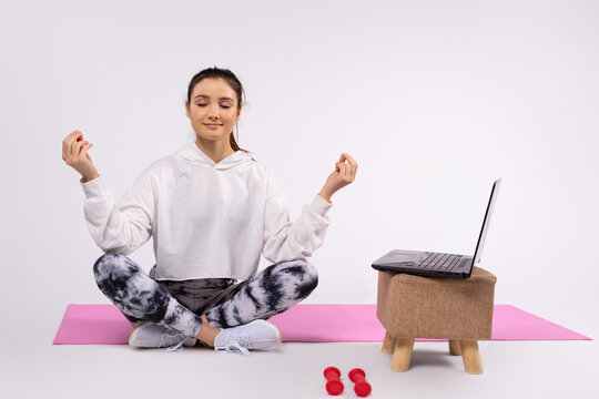 Concentrated Young Girl In Comfortable Clothes While Meditating On A Yoga Mat. Next To A Laptop And Dumbbells. White Background.