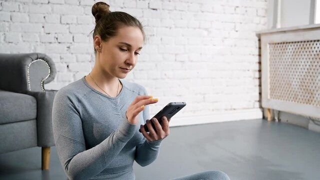 Beautiful woman in on sportswear cloce up, slim woman sit with smartphone. With crossed legs on mat in bright living room .