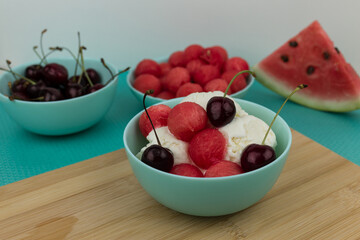 Vanilla ice cream with watermelon balls and cherries in a light turquoise bowl on a bamboo board