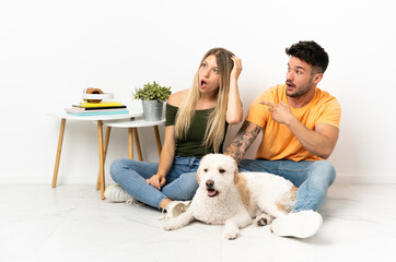 Young caucasian couple with dog staying at home pointing finger to the side with a surprised face