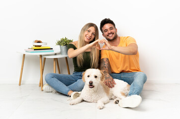 Young caucasian couple with dog staying at home making a heart with hands