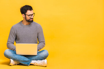 Portrait of a happy young bearded man in casual holding laptop computer while sitting on a floor isolated over yellow background. - 423798122