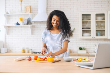 Smiling pretty young African American girl watching cooking classes and learning how to make delicious vegan dinner lunch pizza watching video blog course on laptop in modern kitchen, chopping veggies