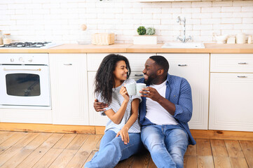 Young mixed-race black couple husband and wife sitting on modern kitchen floor, drinking morning coffee, doing cheers with coffee cups, toasting, celebrating, spending holiday weekend together at home