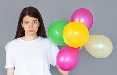 Fototapeta na wymiar Portrait of puzzled upset young woman on pastel gray background with colorful air baloons. Birthday holiday party, people sincere emotions concept
