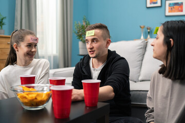 In the Living Room Diverse Group of Fun Loving Friends Playing Who am I game With Sticky Papers Attached to Foreheads. Guys and Girls Trying to Guess What is Written on Sticky Notes