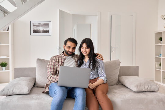 Happy indian family couple using laptop looking at computer sitting on sofa together relaxing at home. Smiling ethnic husband and wife doing online ecommerce shopping on website in modern living room.