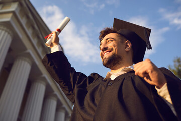 Low angle portrait of happy triumphant male graduate standing near university holding up diploma. From below of young handsome man proud of academic achievements celebrating college graduation - Powered by Adobe