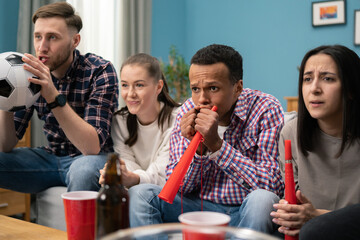 A young African-American male student keeps his fingers crossed for his football team. A group of friends of different nationalities watch the game while sitting on the living room couch at home.