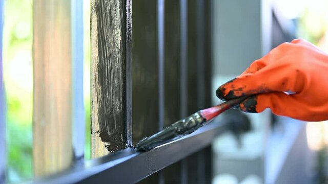 Close up human hand with orange glove holding painting many times on iron fence painting it with bright blue dye. House decorative work concept.