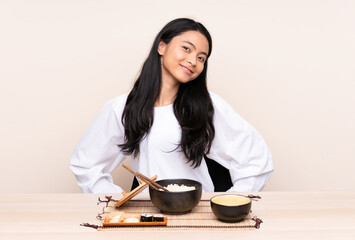 Teenager Asian girl eating asian food isolated on beige background posing with arms at hip and smiling