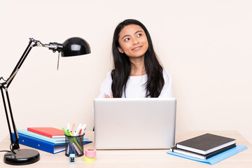 Student asian girl in a workplace with a laptop isolated on beige background looking up while smiling