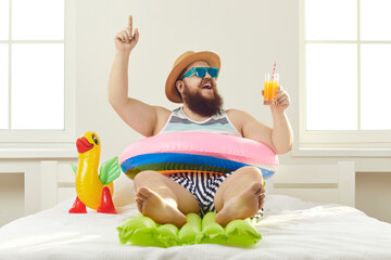 Funny guy in sunglasses, sun hat and swim ring sitting on beach mattress and sipping cocktail...