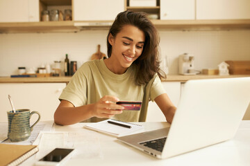 Stylish pretty student girl surfing internet in kitchen, sitting at table with open laptop and mug,...