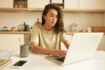 Fototapeta premium Serious young Latin female freelancer keyboarding on portable computer, working all day long, having tired facial expression, drinking another cup of coffee. Remote job, occupation and technology