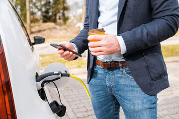Business man standing near the charging electric car , drinking coffee and using his smartphone. Zero emission car. 