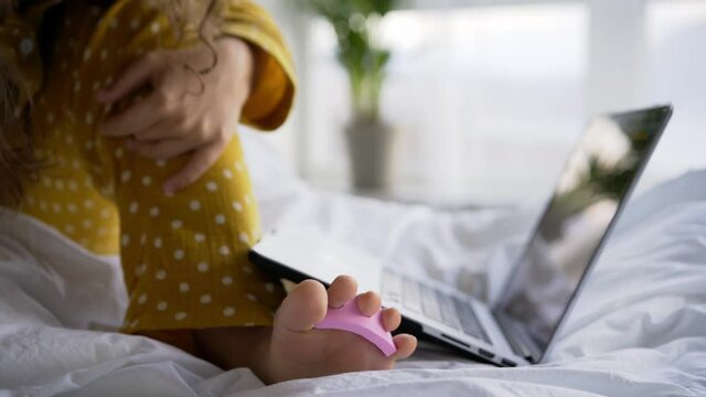 Woman in yellow pajamas puts on pink toe separator and files nail sitting near contemporary laptop on large bed in light room close view crop