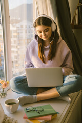 Smiling woman student in headphone study online with laptop at home. Girl sitting near window on...