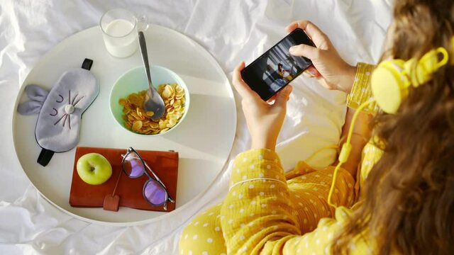 Young woman with headphones looks at pictures on phone near tray with breakfast on comfortable bed in hotel room in lazy morning close upper view