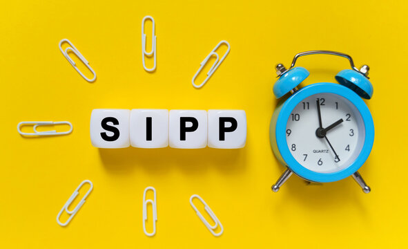 On a yellow background, a blue alarm clock, paper clips and white cubes on which the text is written - SIPP