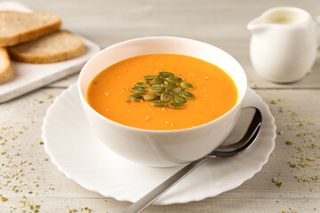 Homemade pumpkin and carrot soup in a white bowl with cream and seeds close up on white wooden rustic background
