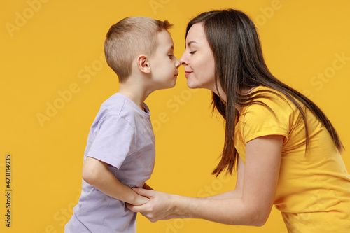 Happy young woman have fun with child baby boy 5-6-7 years old in violet t-shirt touch each other's noses. Mommy little kid son together isolated on yellow background studio. Mother's Day love family.